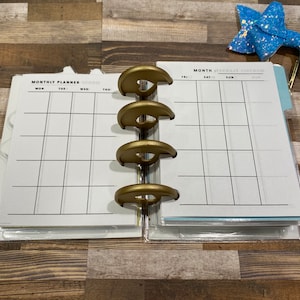 Könnte beinhalten: Ein weißer Monats-Planer mit goldenen Ringen. Der Planer hat zwei Seiten, eine mit der Aufschrift "MONTHLY PLANNER" und die andere mit der Aufschrift "MONTH".