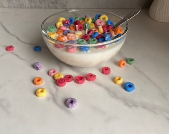 May include: A white bowl filled with colorful cereal rings, resembling a bowl of cereal. The bowl is on a white marble surface with scattered cereal rings.