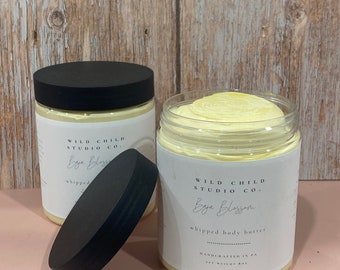 May include: Two glass jars of whipped body butter with black lids. The jars are labeled "WILD CHILD STUDIO CO." and "Baja Blossom" with the text "whipped body butter" and "HANDCRAFTED IN PA".