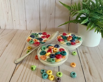 May include: Three small glass bowls filled with white wax and colorful cereal-shaped candles. The bowls are on a light wood surface with scattered cereal-shaped candles.