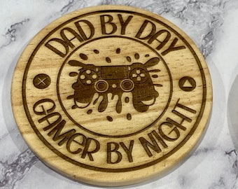 Könnte beinhalten: Ein runder Untersetzer aus Holz mit dem Text "Dad by Day Gamer by Night" und einer Grafik eines Videospielcontrollers mit Spritzfarbe.