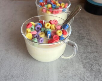 May include: Two clear glass mugs filled with off-white wax that resembles milk, topped with small, colorful, ring-shaped wax pieces in red, yellow, blue, green, and purple. Each mug contains a metal spoon and a central white cotton candle wick.