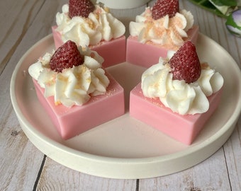 May include: Four pink and white strawberry shortcake-themed soaps on a white plate. Each soap has a red strawberry on top, white frosting, and a sprinkle of pink and white.