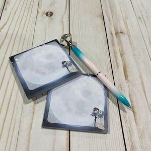 Puede incluir: Dos blocs de notas adhesivas cuadrados con una luna nevada y una pareja que se toma de la mano en cada bloc. Un bolígrafo azul y blanco con una parte superior de cristal está encima de los blocs.