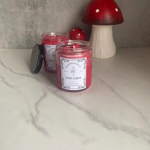 May include: Two red soy wax candles in glass jars with black lids. The candles are labeled "Wild Child Studio Co." and "Cherry Cobbler".