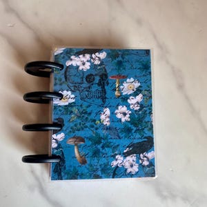May include: A blue and white planner with a skull, mushrooms, and birds on the cover. The planner has a clear plastic cover and a black ring binding.
