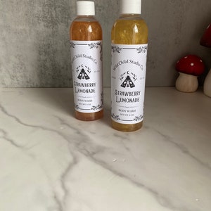 Könnte beinhalten: Zwei bernsteinfarbene Glasflaschen mit goldenem Glitzer. Beide Flaschen haben weiße Etiketten mit schwarzem Text, der "Wild Child Studio Co." und "Strawberry Lemonade" Duschgel lautet. Die Flaschen stehen auf einer weißen Marmorfläche.