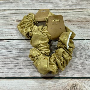 May include: Two gold scrunchies with a metallic gold pattern. Each scrunchie is attached to a brown cardboard tag. The scrunchies are made of a soft, gathered fabric.