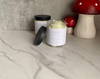 May include: Two jars of whipped body butter with a white label that reads "Wild Child Studio Co. Matcha Whipped Body Butter". The jars are on a white marble surface with a red and white mushroom figurine in the background.