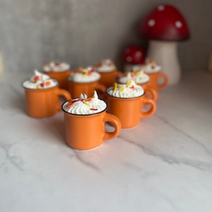 May include: Orange ceramic mugs filled with whipped cream and colorful sprinkles. Each mug has a black rim and a handle. A decorative mushroom is in the background. The mugs are arranged on a white marble surface.