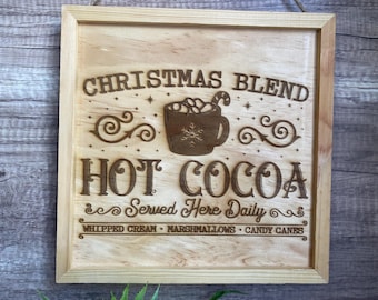 Op de afbeelding: Houten bord met een lichtbruine lijst. Het bord leest "Christmas Blend Hot Cocoa Served Here Daily" in een decoratief lettertype. Een mok cacao met marshmallows en een zuurstok staat in het midden.