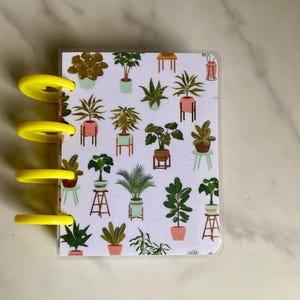 May include: A small, white, plastic-covered notebook with a colorful pattern of potted plants. The notebook has yellow rings on the left side.