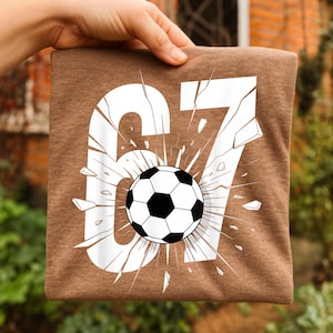 May include: A brown t-shirt with the number "67" in white, and a soccer ball in the center. The number has a cracked effect, with the soccer ball bursting through. The shirt is held up by a hand.