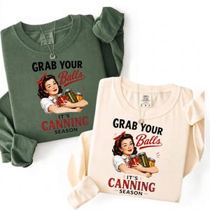 May include: Two long-sleeve crewneck sweatshirts, one in olive green and one in cream, each featuring a graphic of a woman with a red headband holding canning jars. The text reads: GRAB YOUR Balls IT'S CANNING SEASON in black and red lettering.