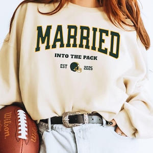May include: A woman wearing a light beige sweatshirt with the text "MARRIED INTO THE PACK EST 2025" and a green football helmet graphic. She is holding a brown football and wearing light blue denim shorts with a brown belt.