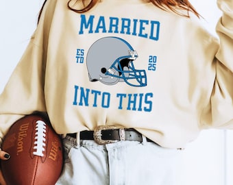May include: A light beige sweatshirt with the text "MARRIED INTO THIS" in blue lettering. A football helmet is centered above the text with the text "ESTD 2025" above the helmet. A woman is wearing the sweatshirt and holding a football.