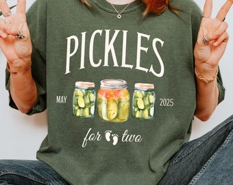 May include: Green crew neck sweatshirt with the text "Pickles for Two" and the date "May 2025". The graphic features three jars of pickles. The person is wearing blue jeans and white sneakers.