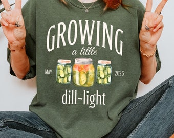 May include: A green t-shirt with the text "GROWING a little dill-light MAY 2025" and a graphic of three jars of pickles.