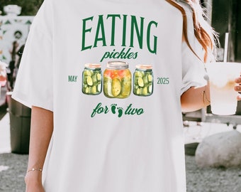 May include: White t-shirt with green text that reads "Eating pickles for two" with a drawing of three jars of pickles and the date "May 2025".
