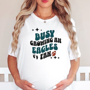 May include: White t-shirt with the text "Busy Growing an Eagles Fan" in teal and black lettering with a football graphic and footprints.