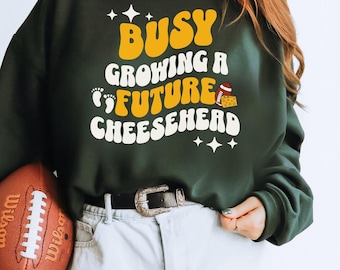 May include: Green sweatshirt with a yellow and white graphic that says "Busy Growing a Future Cheesehead" with a football and cheese graphic. The model is holding a brown football.