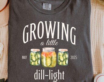 May include: Dark gray t-shirt with the words "GROWING a little dill-light" in white script. The design includes three watercolor jars of pickles, with "MAY 2025" printed on either side. The shirt is folded neatly.