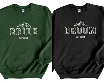 May include: Two sweatshirts, one green and one black, with a mountain range graphic and the words "BRIDE EST 2024" and "GROOM EST 2024" in white.