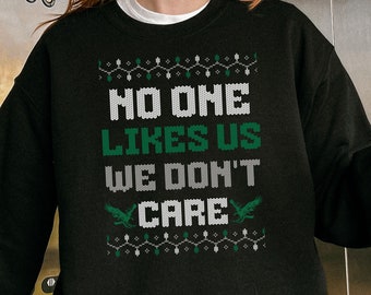 Może przedstawiać: Czarna bluza z napisem "NO ONE LIKES US WE DON'T CARE" w kolorze białym i zielonym. Projekt zawiera elementy dekoracyjne, takie jak lampki choinkowe i dwie zielone postacie przypominające smoki. Wygodny, casualowy element garderoby.