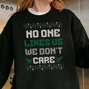 Peut inclure: Sweat-shirt noir avec l'inscription "NO ONE LIKES US WE DON'T CARE" en blanc et vert. Le motif comprend des &eacute;l&eacute;ments d&eacute;coratifs tels que des guirlandes lumineuses et deux figures vertes ressemblant &agrave; des dragons. V&ecirc;tement d&eacute;contract&eacute; et confortable.