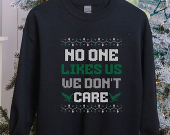 Może przedstawiać: Czarna bluza z napisem "NO ONE LIKES US WE DON'T CARE" w kolorze białym i zielonym. Projekt zawiera dekoracyjne wzory i dwie zielone grafiki ptaków. Bluza wisi na drewnianym wieszaku.