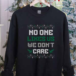 Peut inclure: Sweat-shirt noir avec l'inscription "NO ONE LIKES US WE DON'T CARE" en blanc et vert. Le motif comprend des motifs d&eacute;coratifs et deux graphiques d'oiseaux verts. Le sweat-shirt est suspendu &agrave; un cintre en bois.
