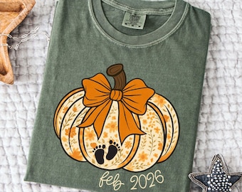 May include: Olive green t-shirt featuring a pumpkin design with an orange bow, floral details, and baby footprints. The text "feb 2026" is below the pumpkin. The shirt is folded and lies on a white textured surface.