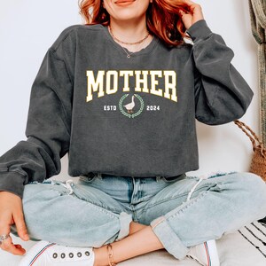 May include: Dark gray sweatshirt with the word "MOTHER" in a yellow collegiate font. Below the word "MOTHER" is a graphic of a goose inside a laurel wreath, with the text "ESTD 2024". The sweatshirt has long sleeves and a crew neck.