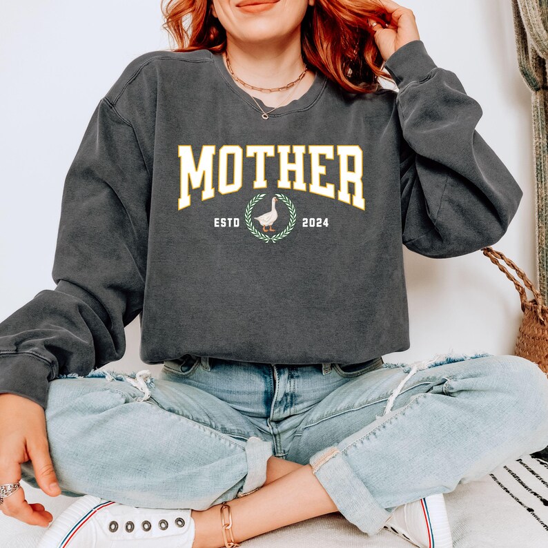 May include: Dark gray sweatshirt with the word "MOTHER" in a yellow collegiate font. Below the word "MOTHER" is a graphic of a goose inside a laurel wreath, with the text "ESTD 2024". The sweatshirt has long sleeves and a crew neck.