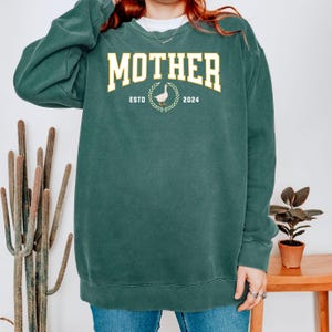 May include: A dark teal sweatshirt with the word "MOTHER" in a yellow collegiate font. Below the word "MOTHER" is a graphic of a goose inside a wreath, with the text "ESTO 2024".