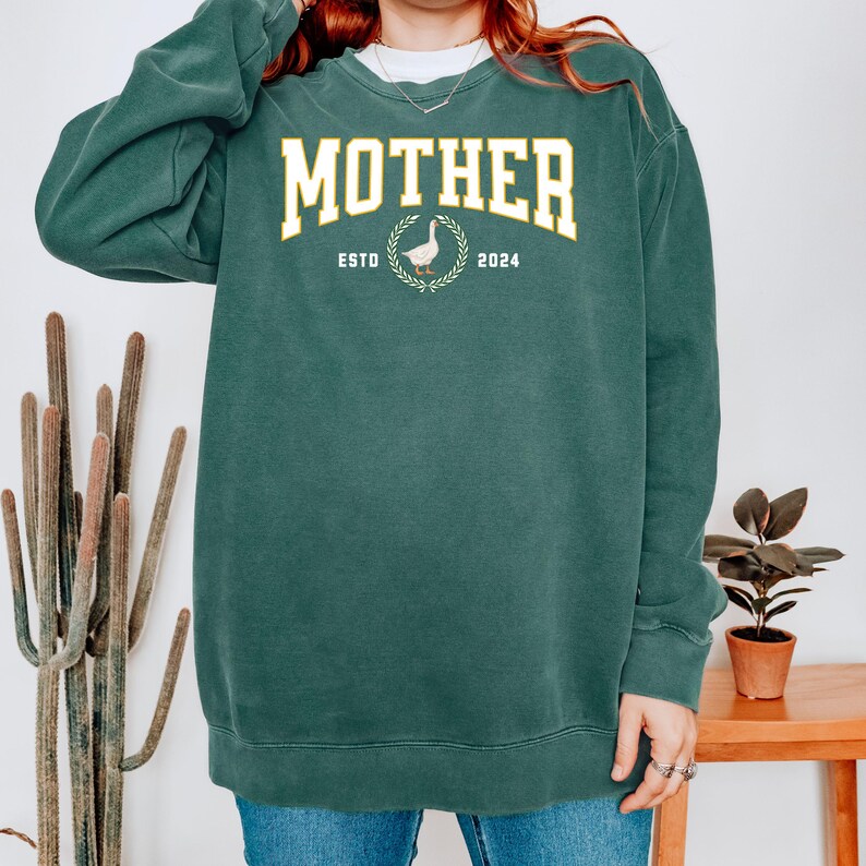 May include: A dark teal sweatshirt with the word "MOTHER" in a yellow collegiate font. Below the word "MOTHER" is a graphic of a goose inside a wreath, with the text "ESTO 2024".