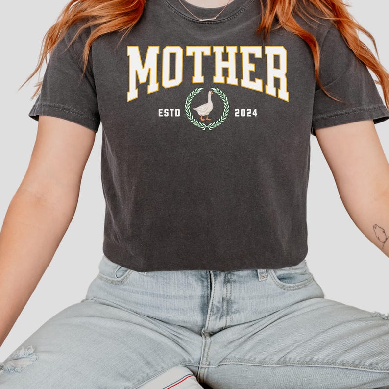 May include: Dark gray t-shirt with the word "MOTHER" in a yellow collegiate font. Below the word is a graphic of a goose inside a laurel wreath, with "ESTO" and "2024" on either side. The shirt is worn by a person.