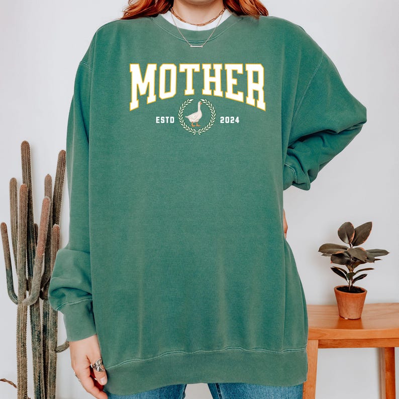May include: Green sweatshirt with the word "MOTHER" in a yellow, collegiate-style font. Below the word "MOTHER" is the text "ESTO 2024" with a goose and wreath graphic in between. The sweatshirt has long sleeves.