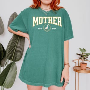 May include: Green t-shirt with the word "MOTHER" in a collegiate font. Below the word "MOTHER" is a goose inside a wreath, with the words "ESTO" and "2024" on either side. The shirt is a solid green color.