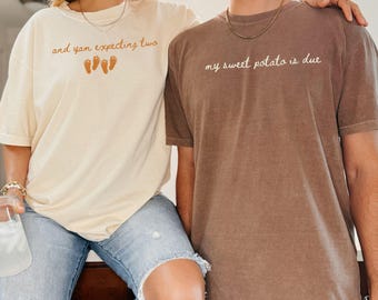 May include: Two matching t-shirts, one cream and one brown, with pregnancy announcement text. The cream shirt says "and yam expecting two" with baby feet graphics. The brown shirt says "my sweet potato is due".
