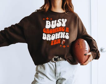 May include: A dark brown sweatshirt with the text "BUSY GROWING BROWNS FAN" in white and orange. The image also includes a brown football with the word "Wilson" printed on it.