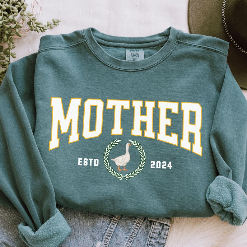 May include: Teal-colored sweatshirt with the word "MOTHER" in large, white, block letters with a gold outline. Below the word is a circular design with a goose illustration and the text "ESTD 2024". The sweatshirt has a crew neck and long sleeves.