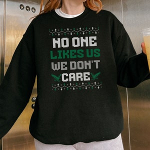 May include: Black sweatshirt with the text "NO ONE LIKES US WE DON'T CARE" in white and green. The design includes two green bird graphics and a string of green and white lights. A casual, festive garment.