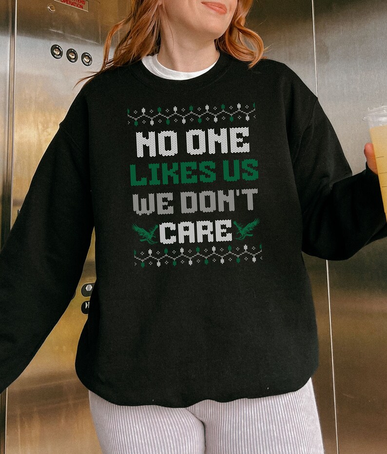 May include: Black sweatshirt with the text "NO ONE LIKES US WE DON'T CARE" in white and green. The design includes two green bird graphics and a string of green and white lights. A casual, festive garment.