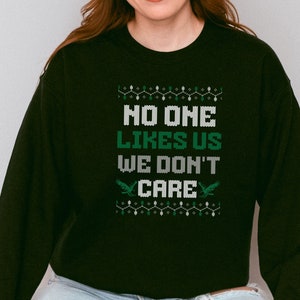 Peut inclure: Sweat-shirt noir avec l'inscription "NO ONE LIKES US WE DON'T CARE" en blanc et vert. Le motif comprend des &eacute;l&eacute;ments d&eacute;coratifs et deux graphiques d'aigles verts. V&ecirc;tement d&eacute;contract&eacute;.