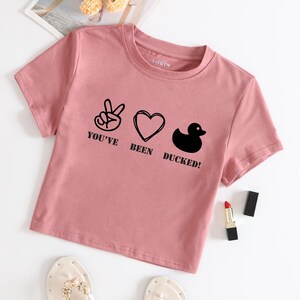 May include: A pink cropped t-shirt with a graphic print of a peace sign, heart, and rubber duck with the text "You've Been Ducked!"