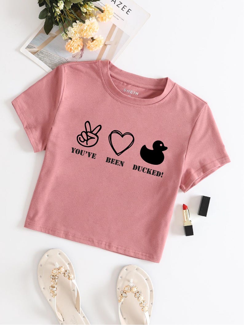 May include: A pink cropped t-shirt with a graphic print of a peace sign, heart, and rubber duck with the text "You've Been Ducked!"