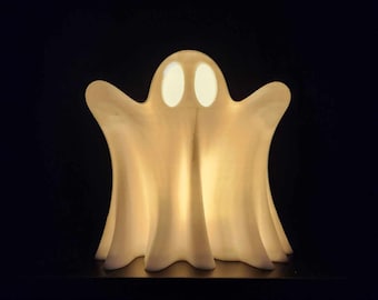 May include: A white ghost-shaped lamp with two glowing eyes. The lamp is lit and casts a warm glow.