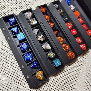 May include: A black folding dice box with multiple compartments for storing dice. The box is open and filled with various colored dice with different numbers.