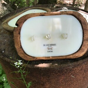 以下が含まれることがあります： 3つの芯を持つ木製のかめ型のキャンドル。キャンドルは白で、前面に「Oak Grove Handmades」と「Ocean Shores」の文字が書かれています。